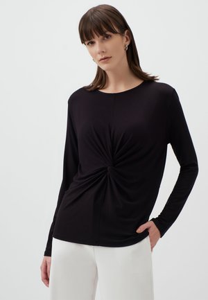 Black long-sleeve top made of soft fabric with a twisted front detail, rounded neckline, and fitted silhouette, paired with white trousers.