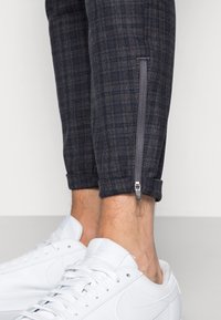 Donkere geruite tapered broeken met een ritsdetail bij de enkel, met een mix van blauw, grijs en subtiele rode lijnen, gecombineerd met witte sneakers.