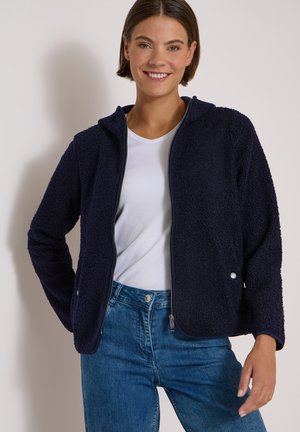 Femme souriante portant une veste en polaire bleu marine zippée par-dessus une chemise blanche et un jean bleu, sur un fond clair uni.