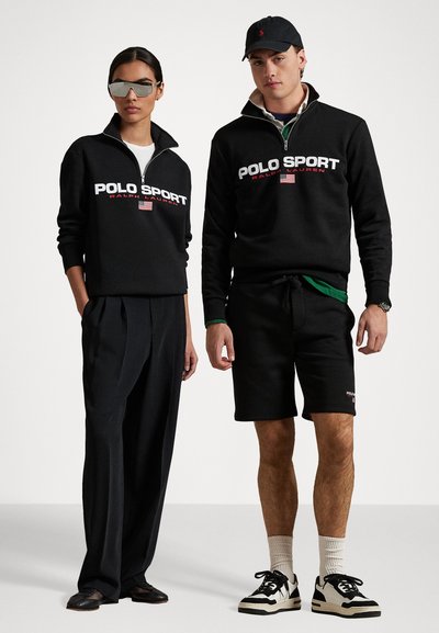 Juodas užtrauktukas su „POLO SPORT“ logotipu, derinamas su juodomis plačiomis kelnėmis. Vyrų modelis juodais šortais ir sportbačiais. Rodyklėje akiniai nuo saulės.