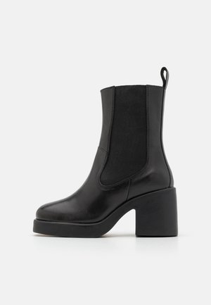 Bottine en cuir noir avec talon bloc, panneau latéral élastique et languette à l'arrière sur un fond gris clair uni.