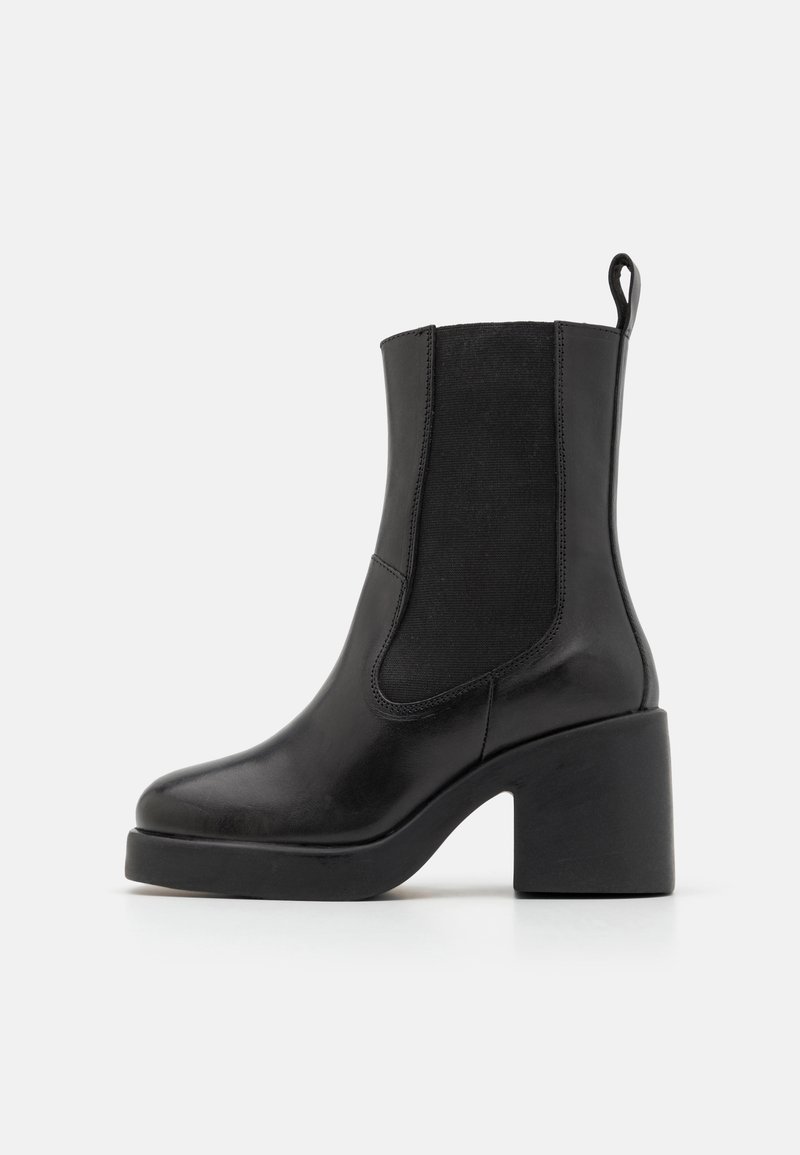 Bottine en cuir noir avec talon bloc, panneau latéral élastique et languette à l'arrière sur un fond gris clair uni.