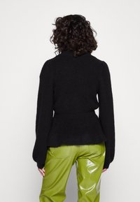 Schwarzer, strukturierter Pullover mit hohem Kragen und Glockenärmeln, kombiniert mit glänzenden grünen Kunstlederhosen. Die Rückansicht zeigt die Designdetails.