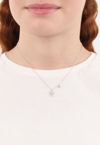 Collana d'argento con un piccolo ciondolo dal design dettagliato e un accento decorativo in oro rosa. Indossata sopra una maglietta bianca.