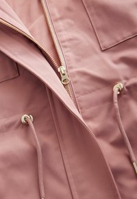Helle rosa Jacke mit einem goldenen Reißverschluss, Zugbändern und zwei Brusttaschen, hergestellt aus einem glatten Stoff mit einer leicht matten Textur.