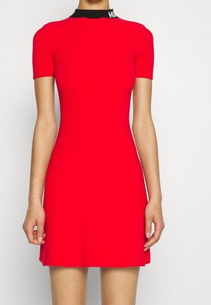 Woman wearing a fitted, short-sleeve red dress with a black mock neck collar featuring white text on the left side.