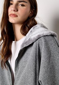 Giacca grigia con zip e cappuccio in pelliccia grigia. Tessuto morbido, vestibilità comoda e con una T-shirt bianca sotto. Primo piano dei dettagli del design.