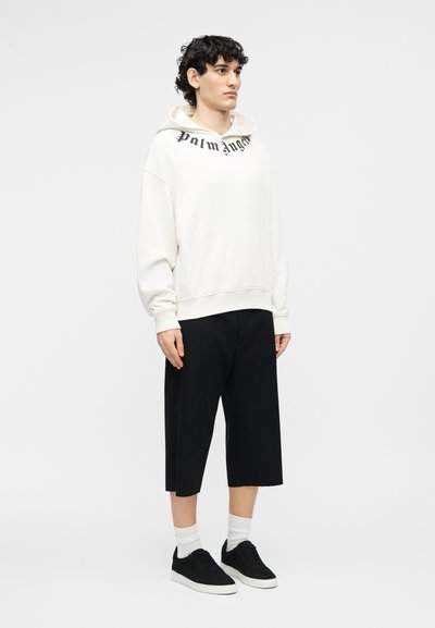 Jeune homme portant un sweat à capuche blanc Palm Angels, un short large noir arrivant au genou, des chaussettes blanches et des baskets noires, debout devant un fond uni.