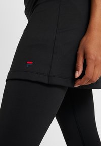 Närbild av en person i svart sportkjol över leggings med en röd och svart logotyp nära fållen, hand vilande vid sidan.
