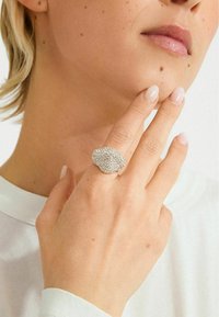 Käsi heleda roosa küünelakiga, millel on suur hõbedane rhinestone rõngas, sõrmed puudutavad lõua, näo osa ja valge särk on nähtavad.