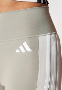 Heledad rohelised legginsid kõrge vöökohtaga, millel on valge kolmerealine disain ja Adidas logo vasakul reiel. Pehme tekstuur.