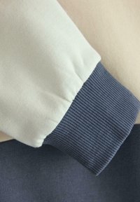 Maglione con corpo beige chiaro, polsini a costine di colore blu scuro a contrasto e texture morbida. Dettaglio di cucitura notevole vicino ai polsini.