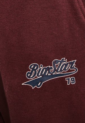 Tumši sarkans svārku krekls ar adītu tekstūru. Tam ir zils logo ar uzrakstu "Big Star" un numuru "79" baltā krāsā.