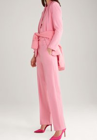 Nainen, joka on pukeutunut vaaleanpunaiseen räätälöityyn bleiseriin, siihen sopiviin korkeavyötäröisiin housuihin ja solmittuun huppariin, yhdistäen kirkkaan vaaleanpunaiset korkokengät.