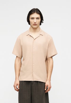 Jeune personne aux cheveux bruns mi-longs portant une chemise beige clair à manches courtes avec des boutons et un pantalon ample foncé, faisant face à l'avant.