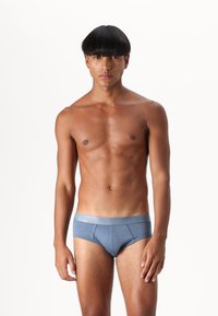 Blå bomullsboxershorts med ett kontrasterande elastiskt midjeband, med en slät yta och minimal sömdesign. Modellen står upprätt.