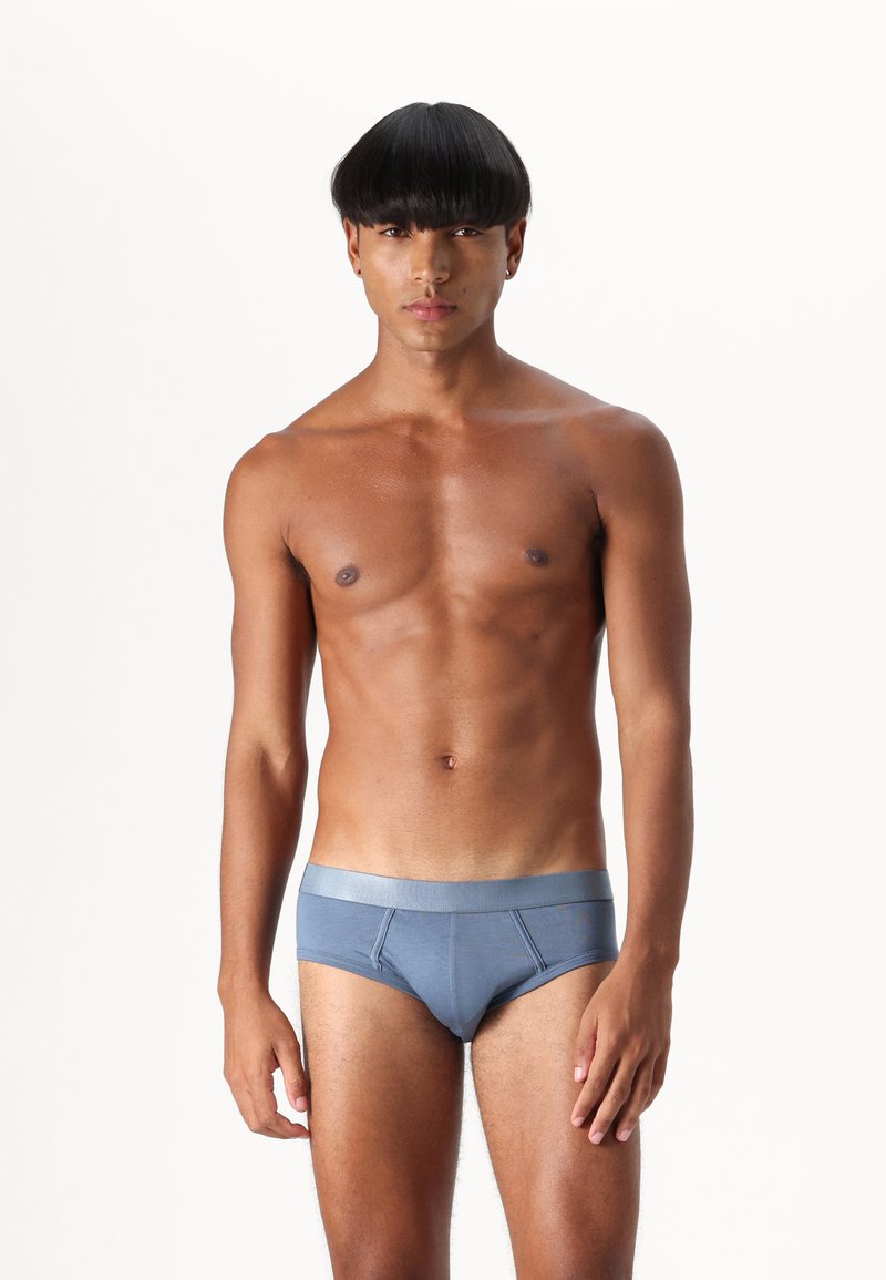 Blå bomullsboxershorts med ett kontrasterande elastiskt midjeband, med en slät yta och minimal sömdesign. Modellen står upprätt.