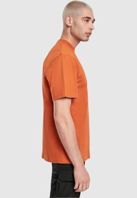 Oranje T-shirt met korte mouwen gemaakt van katoen, met een ronde hals en een relaxte pasvorm. Gemaakt in combinatie met zwarte cargo pants, bekeken vanuit de zijaanzicht.