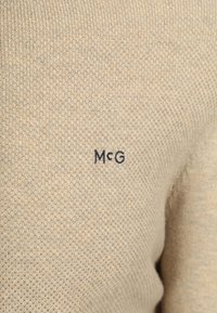 Hellbeige gestrickter Pullover mit einem strukturierten Muster. Auf der Vorderseite befindet sich ein kleines, navyblaues „McG“-Logo. Weicher, mittelschwerer Stoff.