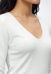 Femme portant un haut blanc à manches longues avec un col en V, montrant l'épaule, le cou et la partie supérieure de la poitrine, avec des cheveux foncés visibles.