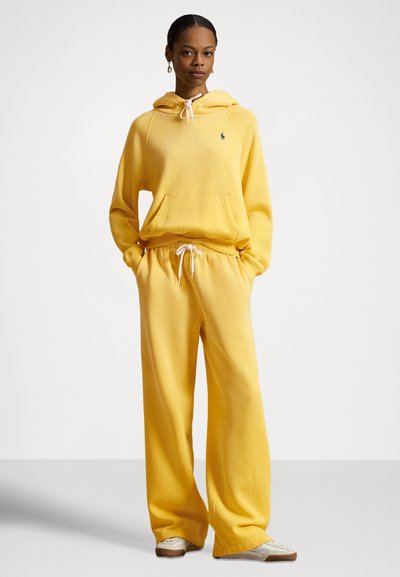 Gul hettegenser og matchende joggebukse laget av mykt stoff. Har hvite snorer, en frontlomme og en liten logo.