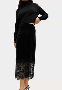 Zwarte fluwelen midi-rok met een kanten zoom met bloemenpatronen. G gedragen met een zwarte longsleeve top en zwarte haksandalen.