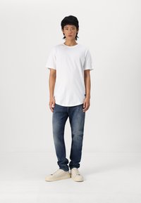 Camiseta blanca de algodón con mangas cortas, combinada con jeans de mezclilla azules. Calzado son zapatillas beige con una suela texturizada.