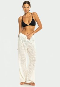 Fekete háromszög bikini felső laza, áttetsző, fehér mintás nadrággal párosítva. A nadrág zigzag mintázatú, laza szabású és könnyű anyagból készült.