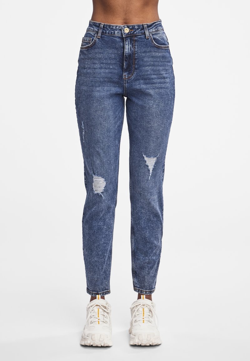 Džínové kalhoty s vysokým pasem v modré barvě, s distressed detaily na kolenou, klasický design s pěti kapsami a slim fit. Kombinované se světle barevnými teniskami.
