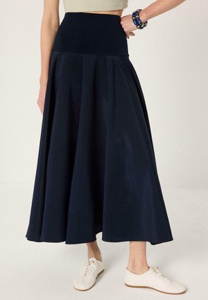 Γυναίκα φοράει μια midi φούστα σε σκούρο μπλε με ψηλή μέση και λευκά παπούτσια με κορδόνια, στέκεται με το ένα χέρι στη μέση και φοράει μπλε βραχιόλι.