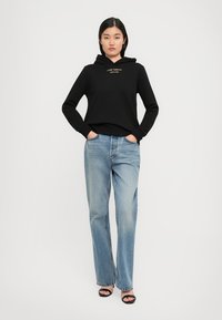 Sweat à capuche noir avec logo doré, associé à un jean droit bleu clair. Tissu texturé et coupe décontractée visibles. Sandales à talons noires.