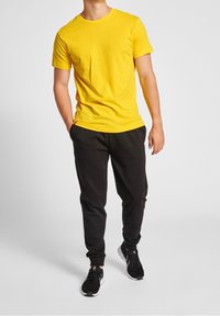 T-shirt in cotone giallo con scollo rotondo, abbinata a pantaloni sportivi neri. Il modello indossa scarpe da ginnastica nere con accenti bianchi.