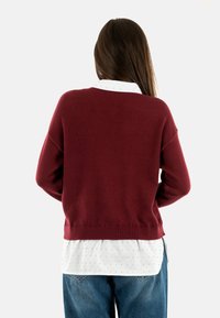 Maglione in maglia bordeaux con vestibilità ampia e orlo a coste, indossato sopra una camicia bianca a pois, abbinato a jeans blu.