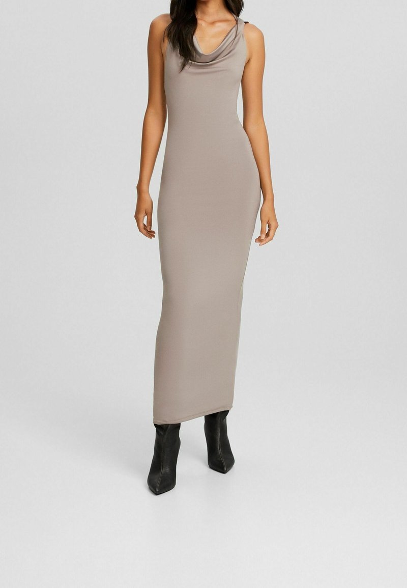 Robe maxi sans manches taupe avec décolleté drapé, assortie à des bottes noires à talons aiguilles, portée par une femme debout.