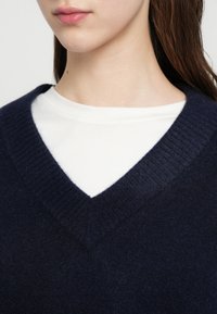 Μπλε σκούρο πουλόβερ με v-λαιμόκοψη και ριμπ πλεκτό τελείωμα, φορεμένο πάνω από λευκό μπλουζάκι με στρόγγυλη λαιμόκοψη. Μαλακή υφή με casual και εφαρμοστή γραμμή.
