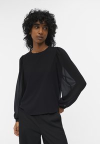 Blusa negra con cuello redondo, tejido suave y mangas abullonadas transparentes con fruncidos en los hombros, combinada con pantalones negros.