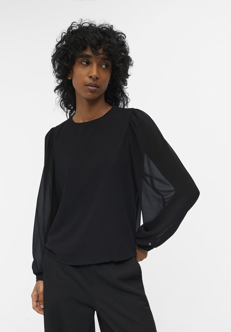 Blusa negra con cuello redondo, tejido suave y mangas abullonadas transparentes con fruncidos en los hombros, combinada con pantalones negros.