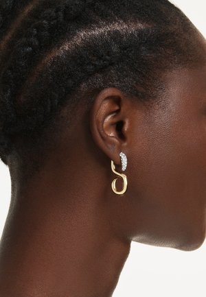Gros plan sur une oreille avec des cheveux noirs tressés, portant une boucle d’oreille dorée de forme abstraite avec une rangée de petites pierres précieuses transparentes.