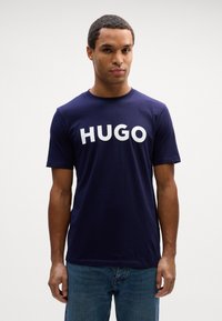 Tums zila T-krekla ar īsām piedurknēm, kas izgatavots no kokvilnas, ar lielu baltu "HUGO" logo krūtīs. Klasisks apaļais kakla dizains.
