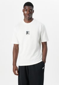 Hvid bomulds t-shirt med et sort og gråt Jordan-logo i et firkantet design på brystet. Model iført med sorte shorts.