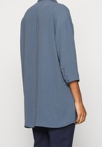 Chemise bleue à manches longues en tissu texturé avec un col à revers, dotée de boutons décoratifs sur les manches et d'une coupe décontractée.