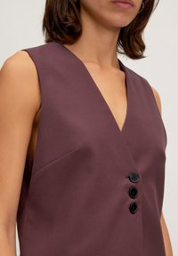 Burgunderfarbenes ärmelloses Top mit tiefem V-Ausschnitt, ausgestattet mit drei schwarzen Knöpfen und einer Seitentasche, aus glattem Stoff gefertigt.