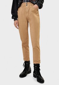 Pantalones de tiro alto color marrón con un ajuste relajado, que cuentan con bolsillos, dos botones frontales y tobillos con puños, combinados con botas negras de cordones.