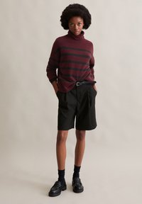 Burgunderfarbener Rollkragenpullover mit schwarzen Streifen, kombiniert mit schwarzen maßgeschneiderten Shorts. Das Modell trägt schwarze Loafers und kniehohe Socken.
