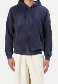 Uomo che indossa una felpa blu navy con zip e logo GAP, abbinata a pantaloni leggeri color beige, in piedi di fronte a uno sfondo neutro.