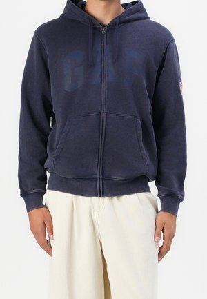 Uomo che indossa una felpa blu navy con zip e logo GAP, abbinata a pantaloni leggeri color beige, in piedi di fronte a uno sfondo neutro.