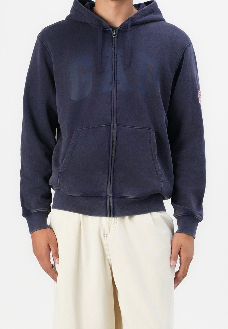 Uomo che indossa una felpa blu navy con zip e logo GAP, abbinata a pantaloni leggeri color beige, in piedi di fronte a uno sfondo neutro.