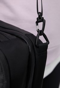 Närbild av svart tygväska rem fäst med plastklämmor och metallfjärilskrok mot en krämfärgad bakgrund.