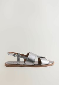 Kovově stříbrné sandály s křížovým designem, nastavitelným páskem kolem kotníku, plochou podrážkou a hnědou stélkou. Minimalistický styl s hladkou texturou.