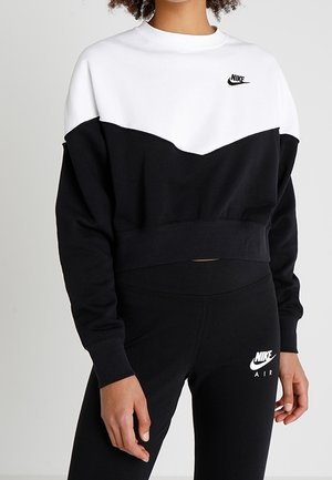 Kobieta ubrana w czarno-białą bluzę Nike oraz czarne legginsy Nike Air, stojąca na tle jednolitego jasnego tła.